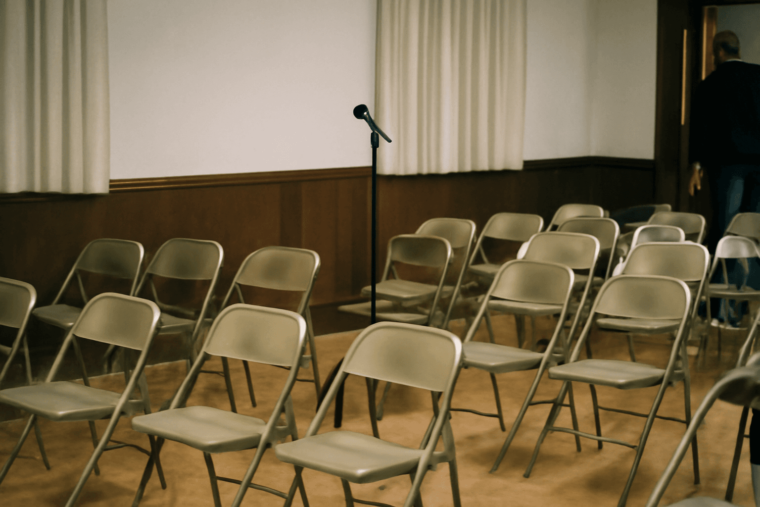 Folding chairs and a microphone stand at the front of a community hall after a crowded evening town hall has emptied out.