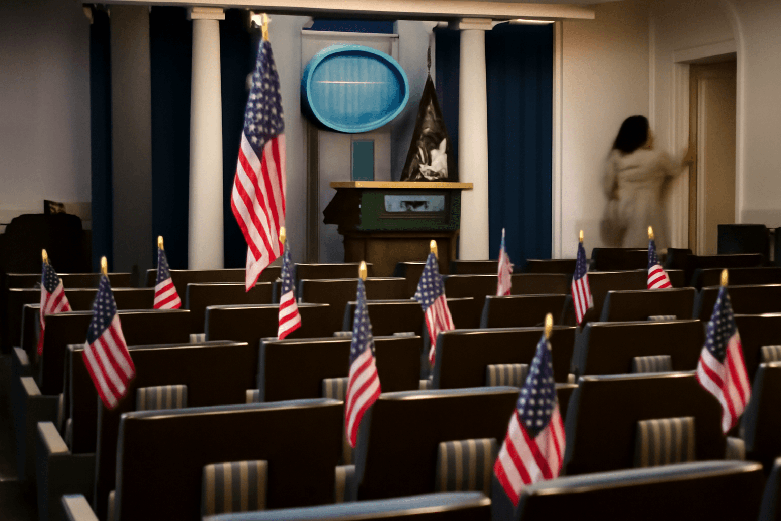 Empty flag-lined chairs in a quiet White House briefing room after a press briefing has ended, seen from the back of the room.